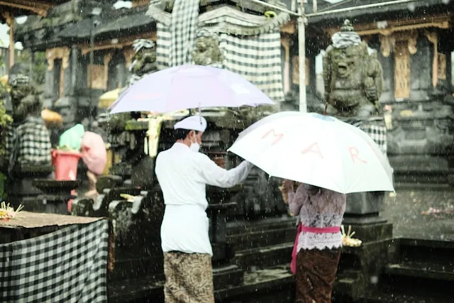 Balinese people holding an umbrella in front of a temple amidst the rain - Weather in Bali