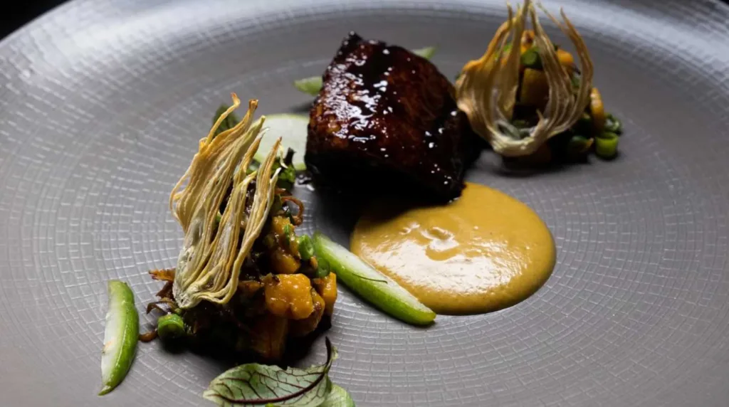A gourmet dish with a glazed protein, artistic arrangements of vegetables, and a smooth sauce, presented on a textured grey plate - best restaurants in seminyak