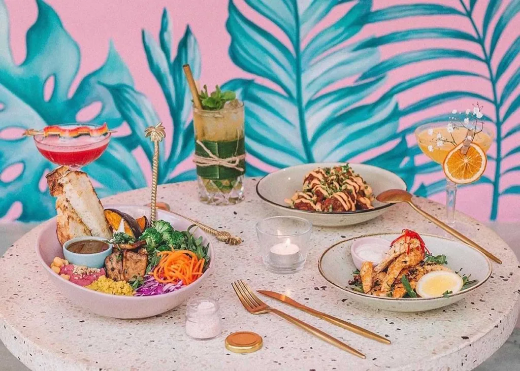Various vibrant dishes and cocktails displayed on a table with a colorful tropical leaf mural in the background