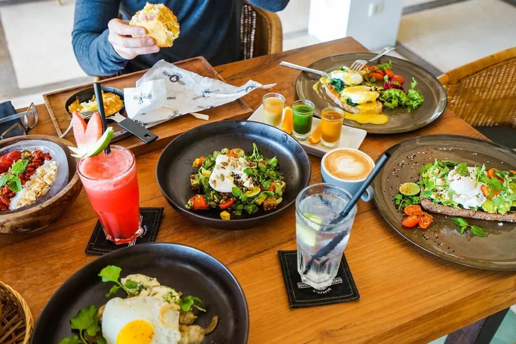 A person dining on a spread of breakfast dishes including avocado toast, oatmeal, salads, a smoothie, and a latte