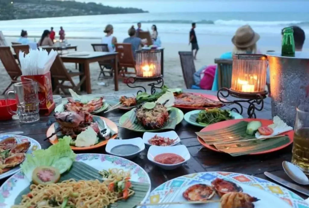 Beachfront dining with a variety of dishes including seafood and noodles on a table, with people and the ocean in the background