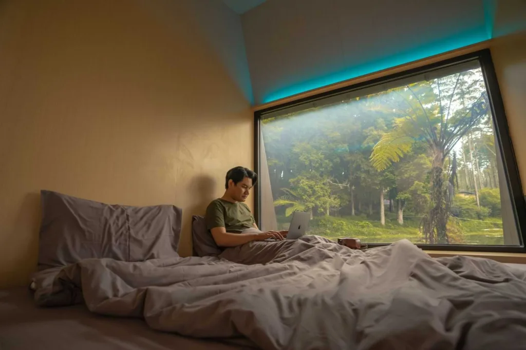 A man using a laptop on a bed beside a large window with a view of lush greenery at Bobocabin
