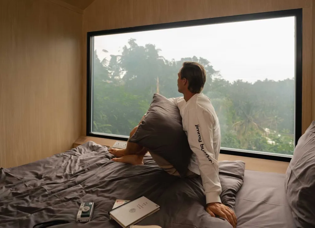 A person sitting in front of large transparent window inside a Bobocabin, staring at the cloudy greenery view outside